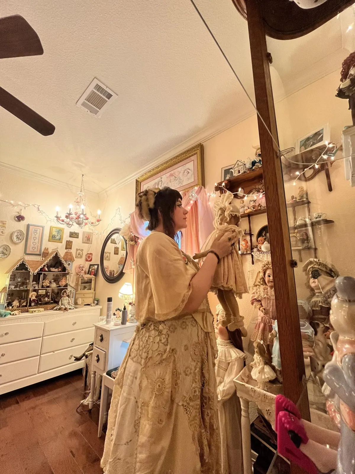 Addilyn Currie in her workroom, lifting an antique doll from a lit cabinet lined with bisque dolls, teacups and a dollhouse.