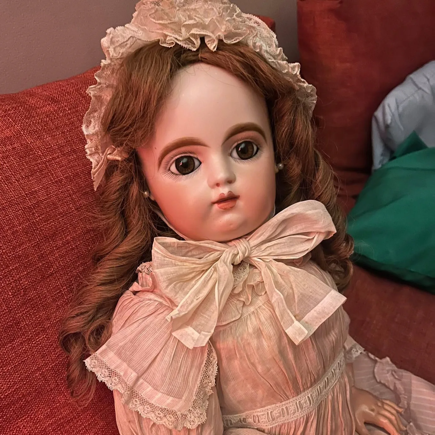 An antique French bébé with auburn mohair curls, brown paperweight eyes and a lace bonnet, in a pale pink dress.