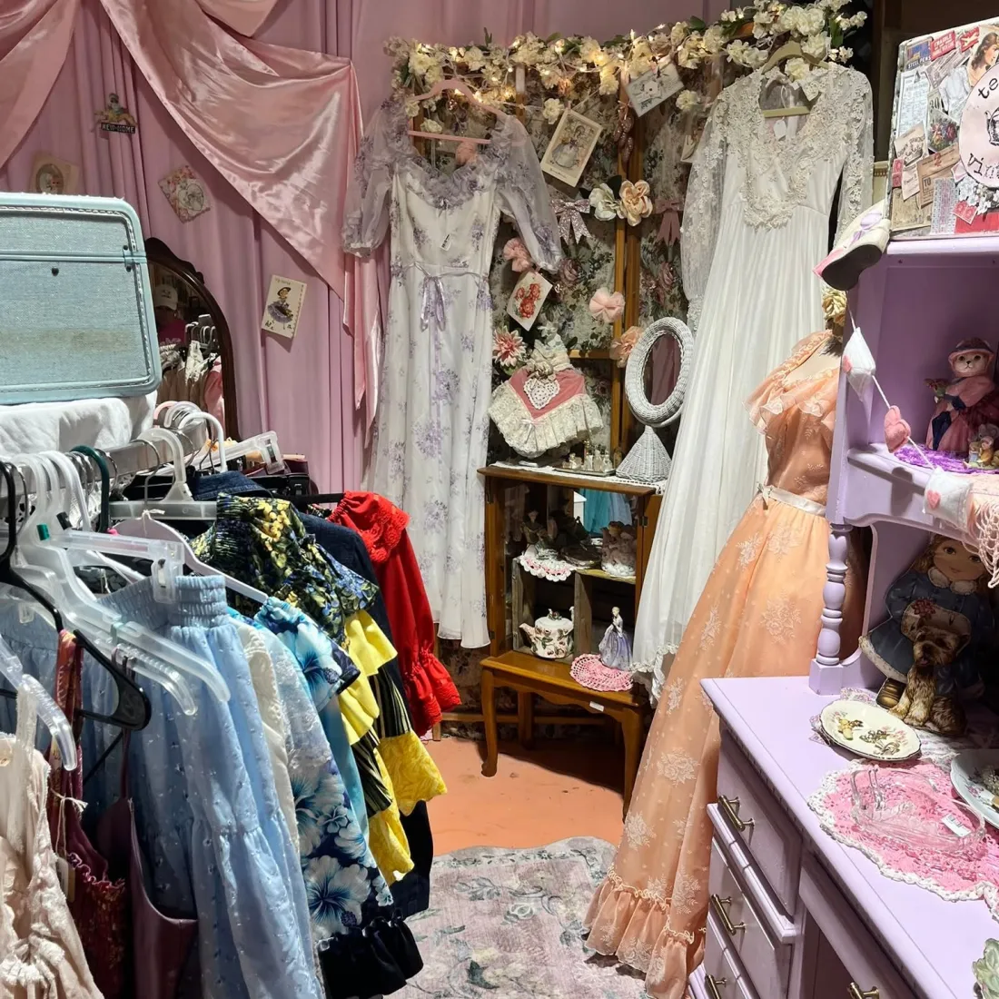 Vintage prairie and lace dresses hanging beside shelves of figurines and a lilac dresser.