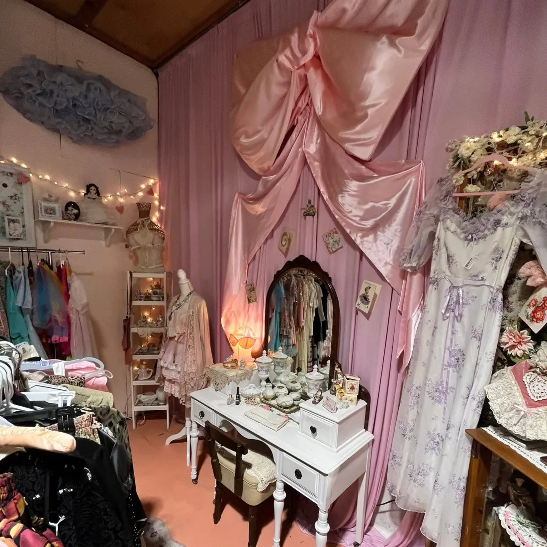 A pink satin-draped corner with a white vanity, mirror, porcelain trinket jars and vintage gowns on a rail.