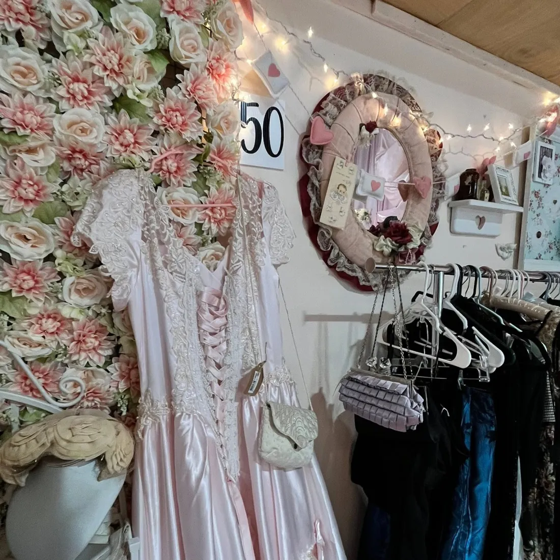 A blush satin and lace gown hanging against a wall of cream and pink silk flowers.