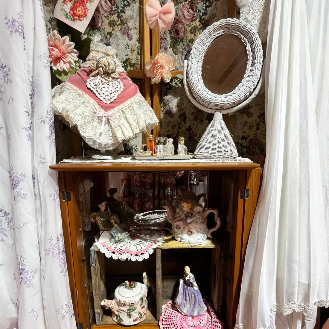 A wooden cabinet holding a floral porcelain teapot, a lady figurine and a lace doll bonnet.